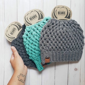 May include: Three handmade messy bun beanies in shades of grey and teal. Each beanie has a textured crochet design and a small leather tag. The beanies are displayed with a cardboard tag that reads "Messy Bun Beanie 100% Handmade."