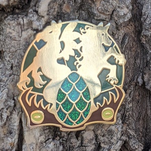 May include: Gold-toned enamel pin featuring three dragons surrounding a green and gold dragon egg. The pin has a detailed design and a shiny finish.