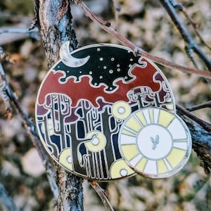May include: A round enamel pin with a black and red forest scene with a crescent moon and stars. The pin also features a yellow sun with a bird flying in front of it.