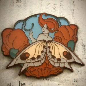 Pode incluir: Um broche de esmalte colorido com uma fada estilizada com cabelo vermelho e asas de borboleta. A fada está cercada por flores de papoula vermelha e um fundo azul.