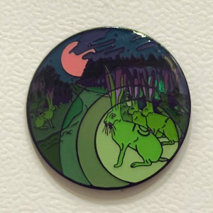 May include: A round enamel pin featuring a nighttime forest scene with green rabbits. The pin has a gradient of green and blue hues, with a pink moon in the sky. The design includes a path and trees.
