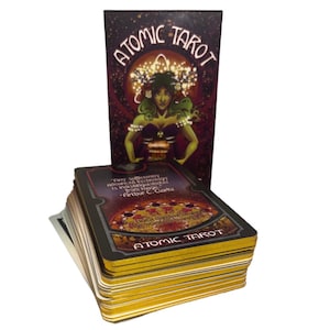 Może przedstawiać: Talia kart Atomic Tarot z pasującym pudełkiem. Pudełko przedstawia kobietę z zieloną sk&oacute;rą i koroną. Karty mają złote krawędzie, a słowa "Atomic Tarot" są wydrukowane na pudełku i kartach.