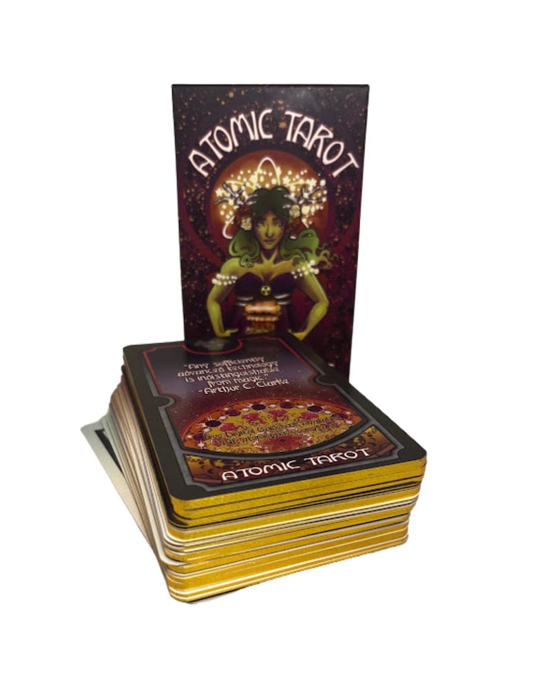 Może przedstawiać: Talia kart Atomic Tarot z pasującym pudełkiem. Pudełko przedstawia kobietę z zieloną sk&oacute;rą i koroną. Karty mają złote krawędzie, a słowa "Atomic Tarot" są wydrukowane na pudełku i kartach.