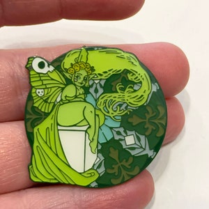 Puede incluir: Un pin de esmalte verde con un hada con alas de mariposa sentada en un pedestal. El hada lleva una corona de flores y tiene el pelo largo y suelto. El fondo es un patrón floral verde y azul.