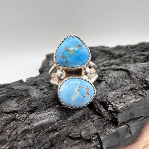 Puede incluir: Un anillo de plata con dos piedras turquesa azules engastadas en un engaste de bisel. El anillo está sobre un fondo de madera oscura.