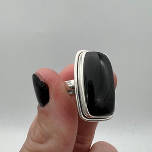 Puede incluir: Un anillo de plata con una piedra rectangular negra. El anillo tiene un estilo simple y moderno.