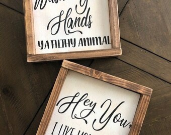 May include: Two square wooden signs with white backgrounds and brown frames. The top sign reads "Wash Your Hands YA FILTHY ANIMAL" in black script. The bottom sign says "Hey You, I LIKE YOUR FACE" in black script.