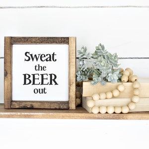 May include: A wooden shelf with black metal brackets, holding a framed sign that reads "Sweat the BEER out." The shelf also displays faux greenery, wooden beads, and two books. The background is a white shiplap wall.