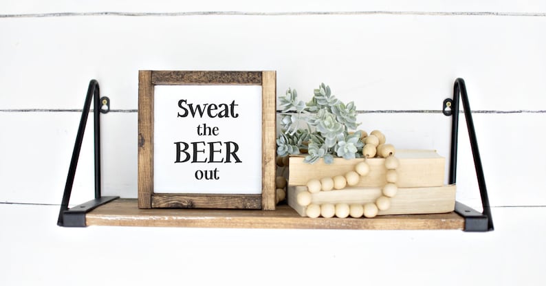 May include: A wooden shelf with black metal brackets, holding a framed sign that reads "Sweat the BEER out." The shelf also displays faux greenery, wooden beads, and two books. The background is a white shiplap wall.