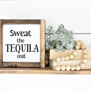 May include: A wooden shelf with black metal brackets displays a framed sign with the text "Sweat the TEQUILA out." The shelf also features artificial plants, wooden beads, and two books. The background is a white shiplap wall.