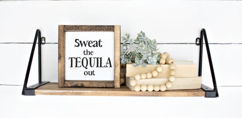 May include: A wooden shelf with black metal brackets displays a framed sign with the text "Sweat the TEQUILA out." The shelf also features artificial plants, wooden beads, and two books. The background is a white shiplap wall.