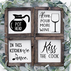 May include: Four framed wooden signs with white backgrounds and black text. The signs read: "Pot Head", "Alexa, Pour More Wine", "In This Kitchen We Dance", and "Kiss the Cook". The signs are surrounded by greenery.