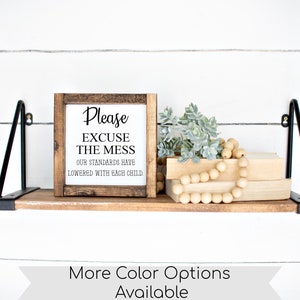 May include: A wooden shelf with black metal brackets holds a framed sign that reads "Please EXCUSE THE MESS OUR STANDARDS HAVE LOWERED WITH EACH CHILD." Decorative items include faux succulents, wooden beads, and books. The shelf is against a white shiplap wall.