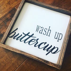 May include: A square wooden sign with a white background and a dark brown frame. The words "wash up buttercup" are written in black cursive script.