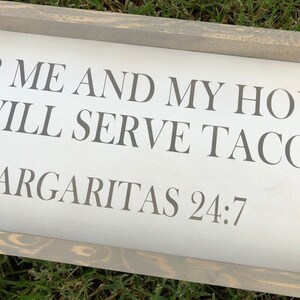 May include: A rectangular wooden sign with a gray frame and white background. The sign features the text "FOR ME AND MY HOUSE WE WILL SERVE TACOS. MARGARITAS 24:7" in gray lettering.