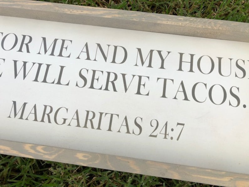 May include: A rectangular wooden sign with a gray frame and white background. The sign features the text "FOR ME AND MY HOUSE WE WILL SERVE TACOS. MARGARITAS 24:7" in gray lettering.