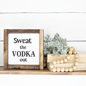 May include: A wooden shelf with black metal brackets displays a framed sign saying "Sweat the VODKA out." The shelf also features faux greenery, wooden beads, and two stacked books. The background is a white shiplap wall.
