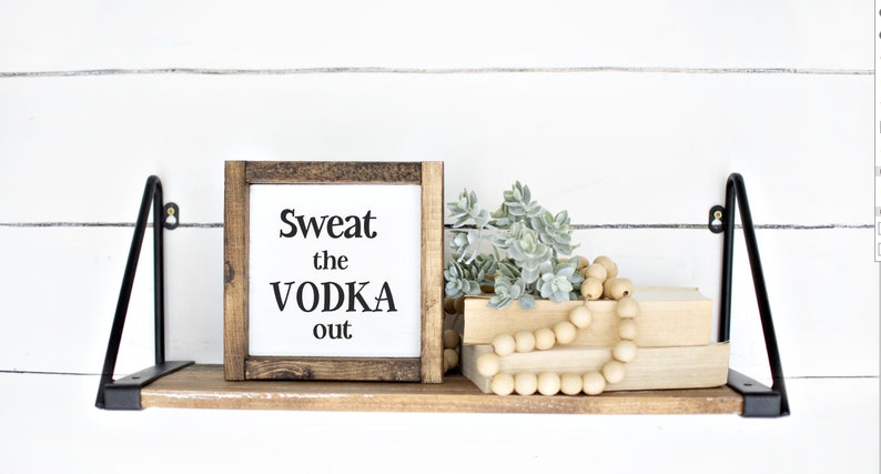 May include: A wooden shelf with black metal brackets displays a framed sign saying "Sweat the VODKA out." The shelf also features faux greenery, wooden beads, and two stacked books. The background is a white shiplap wall.