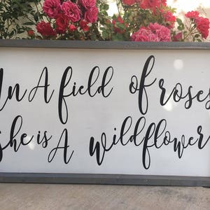 May include: A rectangular wooden sign with a gray frame and a white background. The sign features black cursive text that reads, "In A field of roses she is A wildflower."