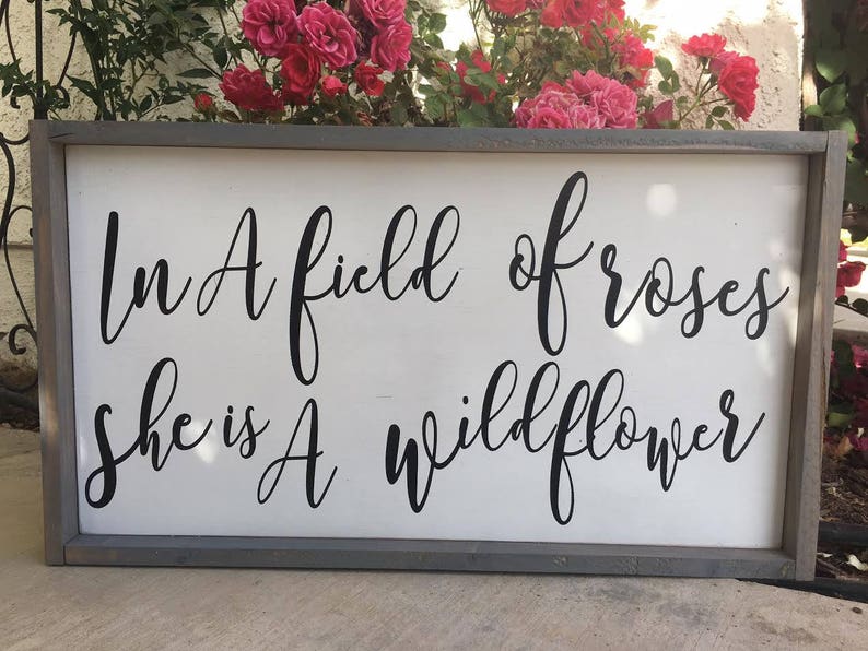 May include: A rectangular wooden sign with a gray frame and a white background. The sign features black cursive text that reads, "In A field of roses she is A wildflower."