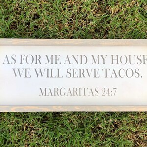 May include: A rectangular wooden sign with a light gray frame, resting on green grass. The sign features the text "AS FOR ME AND MY HOUSE, WE WILL SERVE TACOS. MARGARITAS 24:7" in gray lettering, perfect for a kitchen or dining area.
