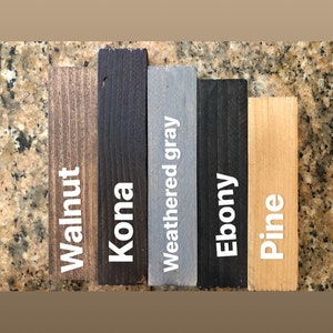 May include: A collection of stained wood samples labeled "Walnut", "Kona", "Weathered gray", "Ebony", and "Pine". The samples are rectangular and vary in color from light brown to black, with the text in white.