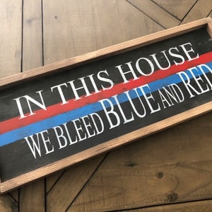 May include: Rectangular wooden sign with a black background and a brown frame. The sign features the text "IN THIS HOUSE WE BLEED BLUE AND RED" in white lettering, with red and blue stripes. The sign is displayed on a wooden surface.