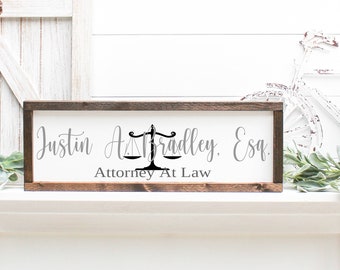 May include: A rectangular wooden sign with a white background and dark brown frame. The sign displays the name "Justin A. Bradley, Esq." in gray script, with a black scales of justice symbol. Below, it reads "Attorney At Law."