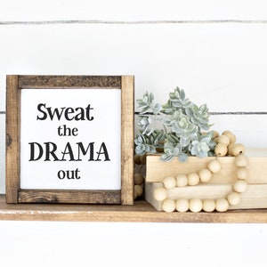 May include: A wooden shelf with black metal brackets displays a framed sign saying "Sweat the DRAMA out." The shelf also features books, faux succulents, and a wooden bead garland. The background is a white shiplap wall.