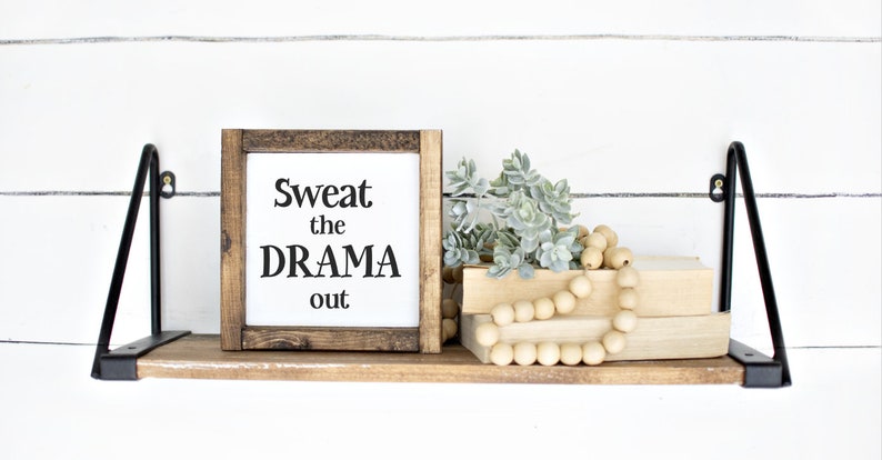 May include: A wooden shelf with black metal brackets displays a framed sign saying "Sweat the DRAMA out." The shelf also features books, faux succulents, and a wooden bead garland. The background is a white shiplap wall.