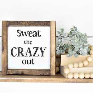 May include: A wooden shelf with black metal brackets displays a framed sign saying "Sweat the CRAZY out." The shelf also features faux succulents, wooden beads, and two stacked books. The background is a white shiplap wall.