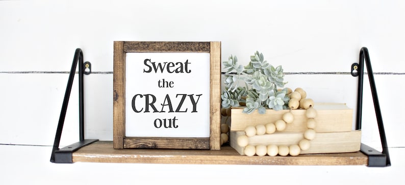 May include: A wooden shelf with black metal brackets displays a framed sign saying "Sweat the CRAZY out." The shelf also features faux succulents, wooden beads, and two stacked books. The background is a white shiplap wall.