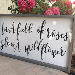 May include: A rectangular wooden sign with a gray frame and a white background. The sign features black cursive text that reads, "In A field of roses she is A wildflower." Pink roses are visible in the background.