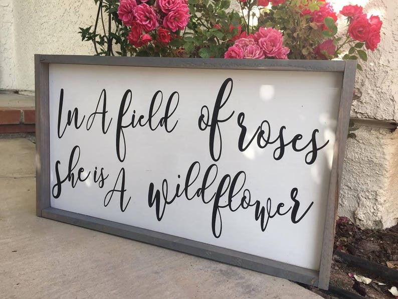 May include: A rectangular wooden sign with a gray frame and a white background. The sign features black cursive text that reads, "In A field of roses she is A wildflower." Pink roses are visible in the background.