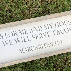 May include: A rectangular wooden sign with a gray frame and white background. The sign reads "AS FOR ME AND MY HOUSE, WE WILL SERVE TACOS. MARGARITAS 24:7" in gray lettering. The sign is placed on a bed of green grass.