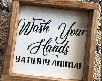 May include: A framed wooden sign with the text "Wash Your Hands Ya Filthy Animal" in black script on a white background. The frame is made of light brown wood.
