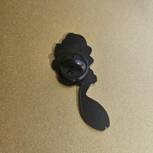 May include: The reverse side of a black metal enamel pin featuring a textured matte finish. A circular black rubber clutch is attached to the center post, which is used to secure the pin to fabric or other materials. The shape is irregular.