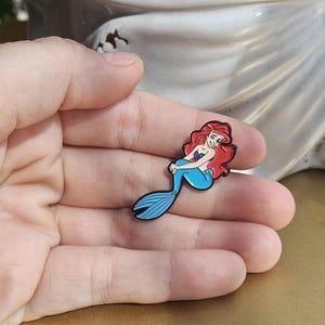 May include: A small enamel pin featuring a cartoon mermaid with long red hair, a purple seashell top, and a bright blue tail. The character is depicted in a seated pose with a smiling expression, outlined in black metal, and finished with smooth enamel.