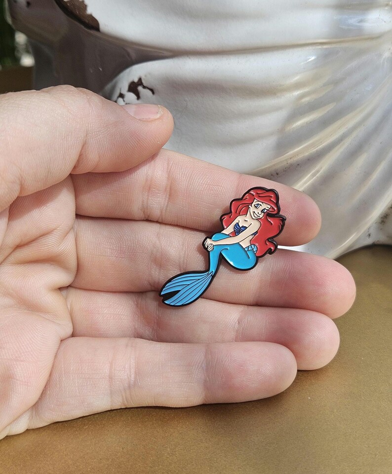 May include: A small enamel pin featuring a cartoon mermaid with long red hair, a purple seashell top, and a bright blue tail. The character is depicted in a seated pose with a smiling expression, outlined in black metal, and finished with smooth enamel.
