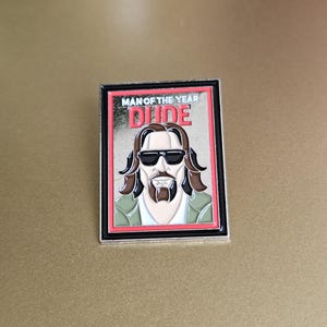 May include: A silver metal pin with a red and black border. The pin features a cartoon image of a man with sunglasses and a beard. The text "MAN OF THE YEAR DUDE" is printed in white on a red background.