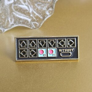 May include: A rectangular enamel pin featuring a retro video game controller design. The pin has a black background with white directional arrows, pink and blue buttons, and the word "START". The pin is set against a gold background.