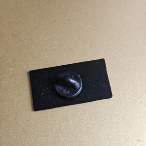 May include: The back of a black enamel pin with a black metal pin back.