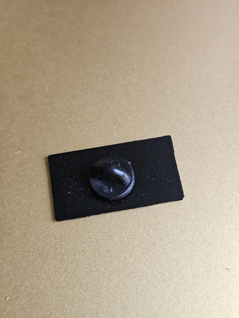 May include: The back of a black enamel pin with a black metal pin back.