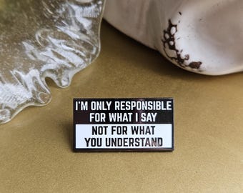 May include: A rectangular enamel pin with the text "I'M ONLY RESPONSIBLE FOR WHAT I SAY NOT FOR WHAT YOU UNDERSTAND" in black and white. The pin is on a gold surface, with a clear glass sculpture and a white ceramic object in the background.