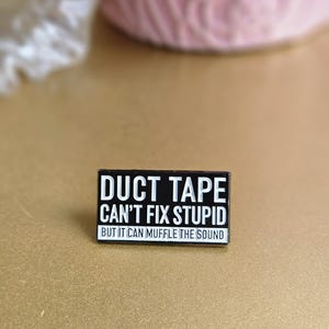 May include: Black enamel pin with white text that reads "Duct Tape Can't Fix Stupid But It Can Muffle The Sound".