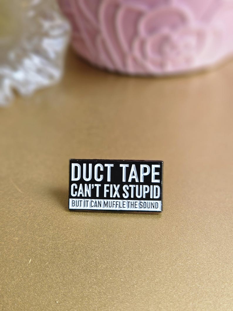 May include: Black enamel pin with white text that reads "Duct Tape Can't Fix Stupid But It Can Muffle The Sound".