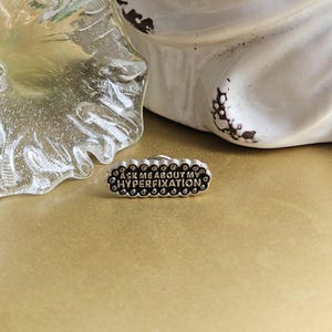 May include: A silver-colored pin with the text "ASK ME ABOUT MY HYPERFIXATION" in black lettering. The pin has a scalloped edge and is set against a gold background. A white ceramic object is in the background.
