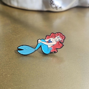 May include: A metal enamel pin featuring a cartoon mermaid with long flowing red hair, a blue tail, and a purple seashell top. The character is depicted in a reclining pose with a smiling expression, outlined in black metal with vibrant colors.