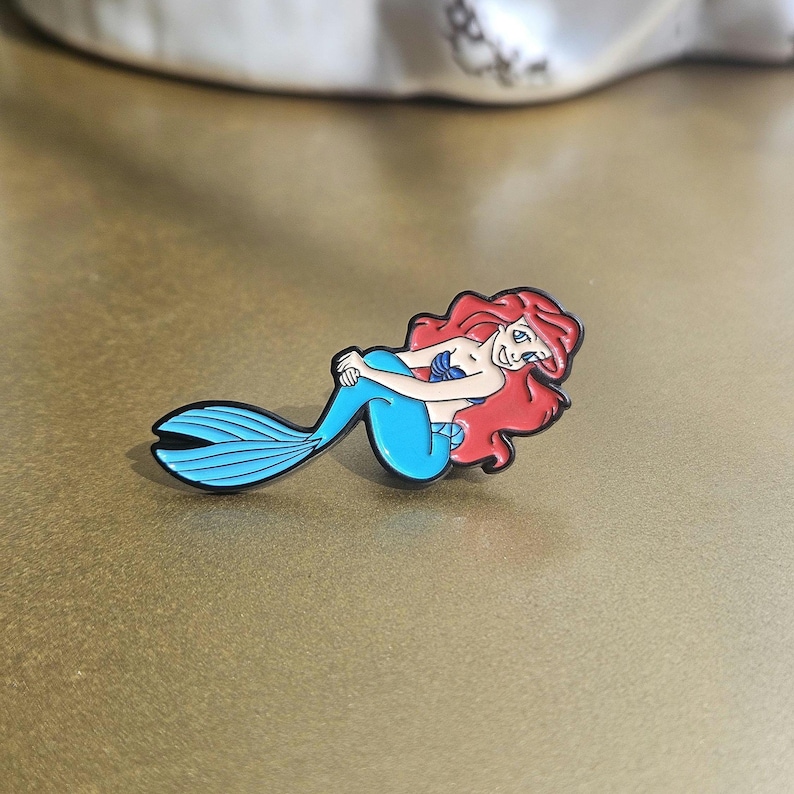 May include: A metal enamel pin featuring a cartoon mermaid with long flowing red hair, a blue tail, and a purple seashell top. The character is depicted in a reclining pose with a smiling expression, outlined in black metal with vibrant colors.