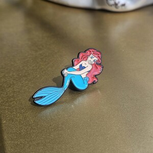 May include: A metal enamel pin featuring a cartoon mermaid with long red hair, a blue seashell top, and a bright blue tail. The character is depicted in a seated pose with a black outline, showcasing vibrant colors and a smooth, glossy finish.
